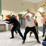 A group of people dancing. Two men are featured mainly, they are stretching their arms out as they do a powerful movement