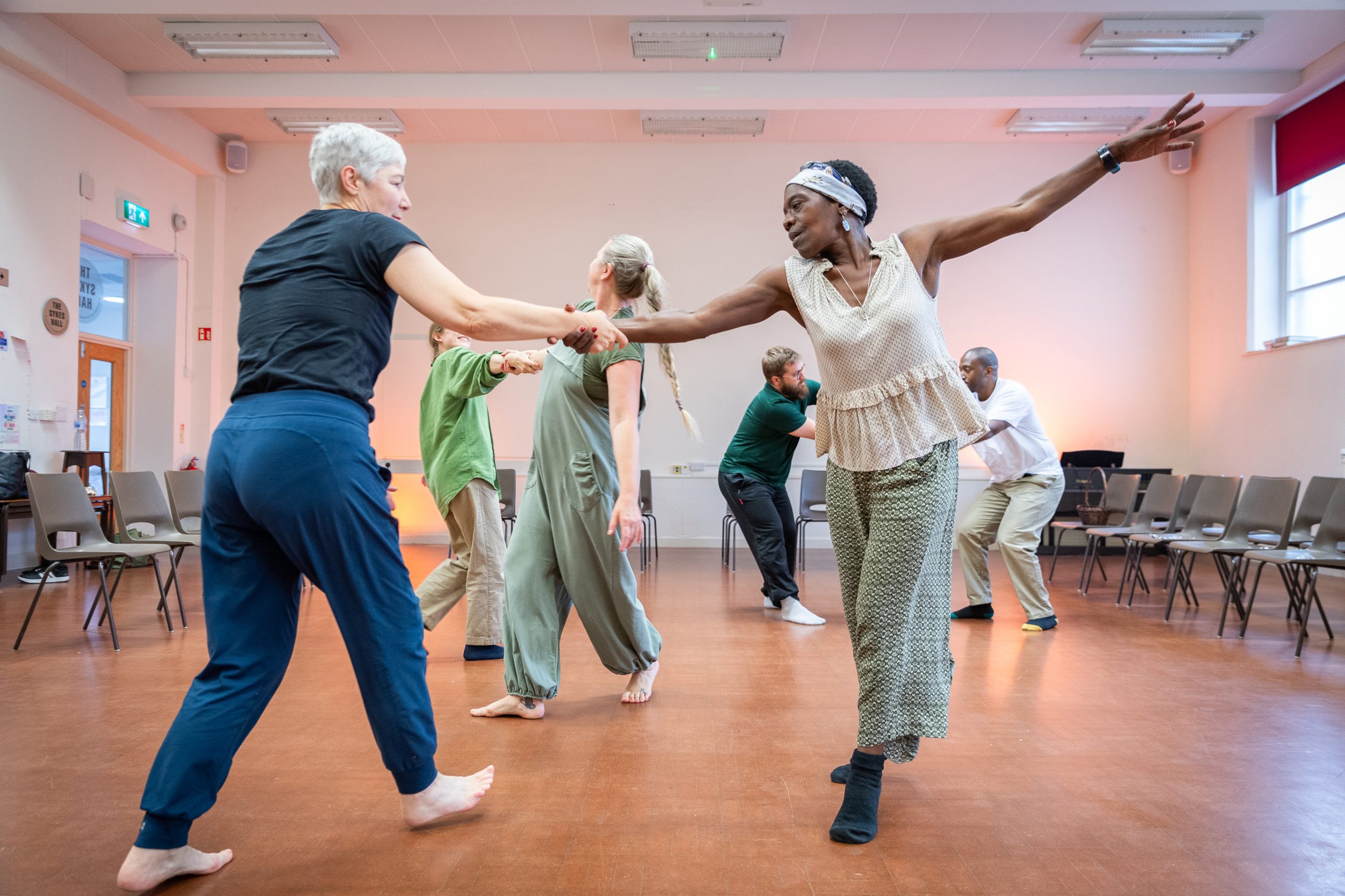 A group of dancers dance in pairs, holding arms as if swinging around the room. Two dancers can be seen in the foreground - one has an arm reaching out wide into the space as they connect with their partner.